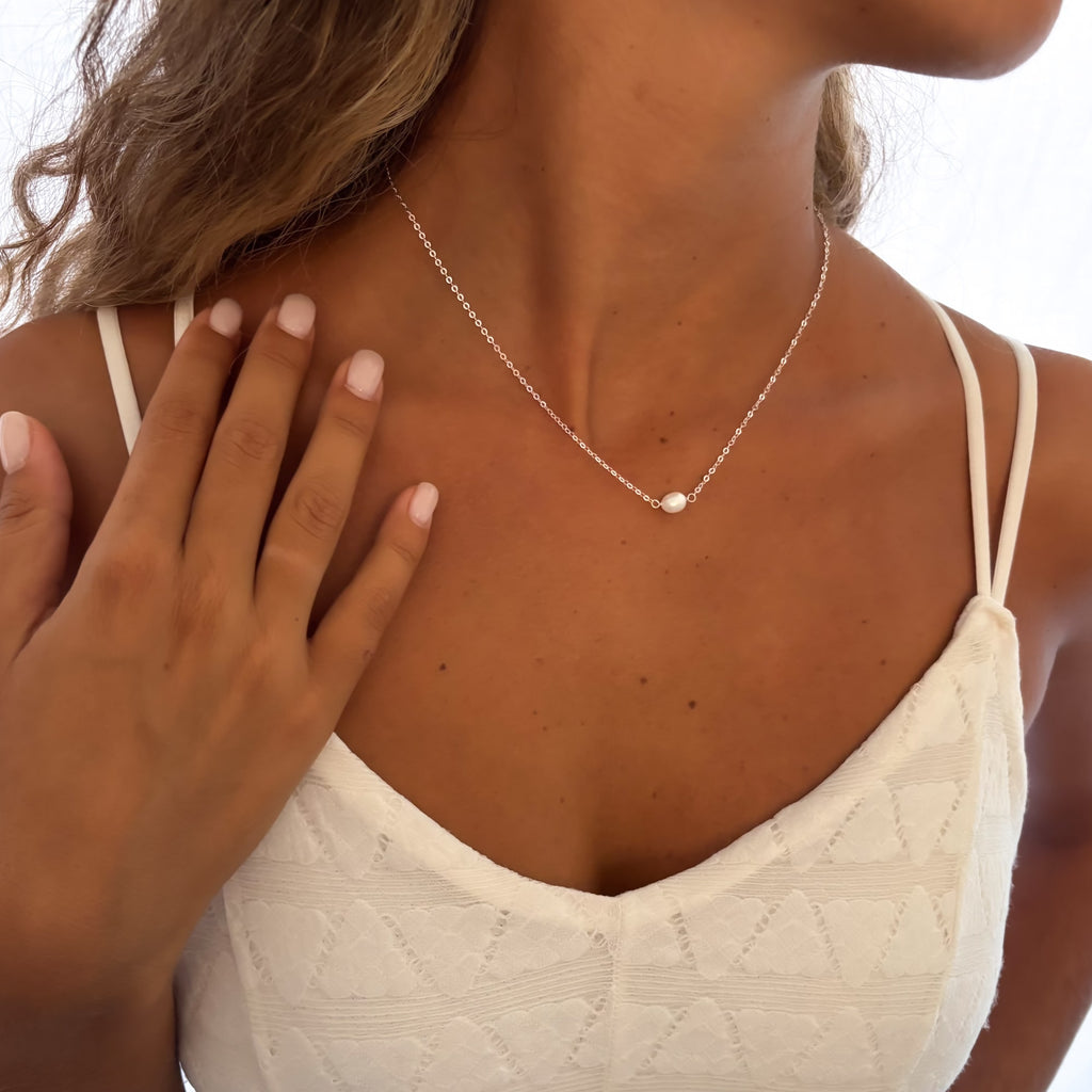 Woman wearing a delicate necklace with a pearl pendant, hand touching her shoulder.