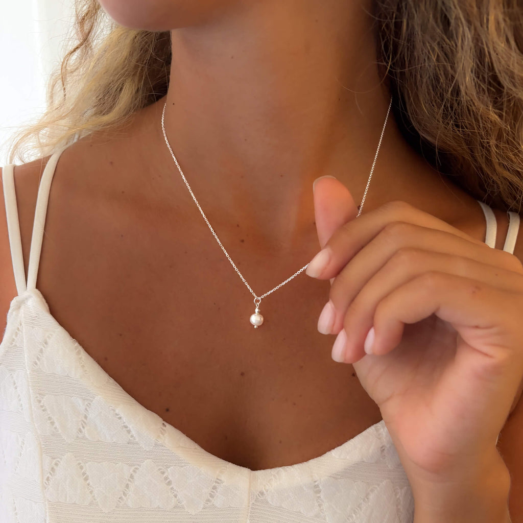 Woman wearing a delicate necklace with a small pearl pendant, close-up of the necklace and hand holding the dainty silver chain.