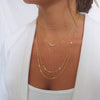 Model wearing layered necklaces. all necklaces are gold. The necklace stack is made from a tube chain choker , two charm necklaces and long chain necklaces